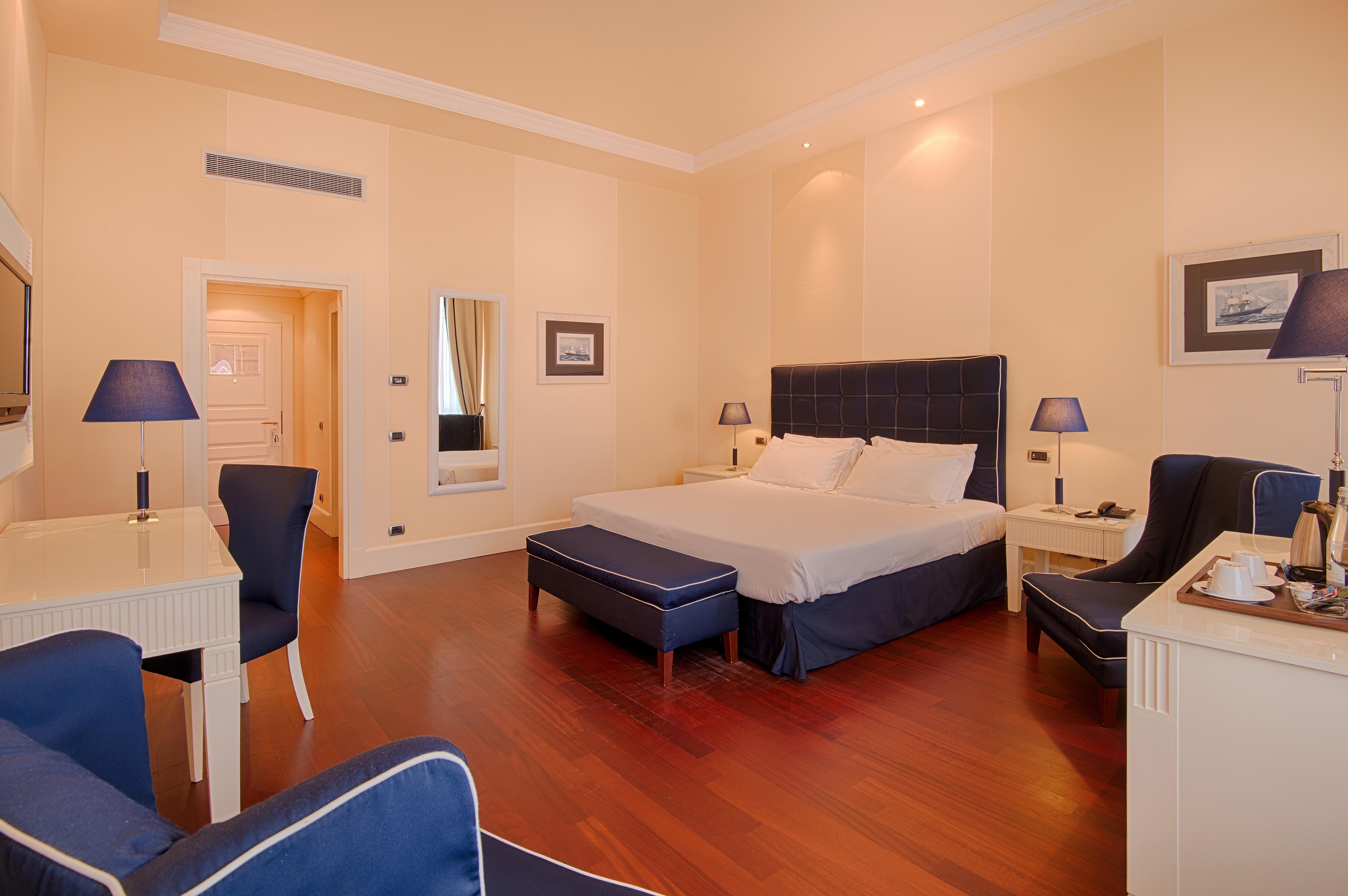 Grand Hotel Palazzo Livorno MGallery Collection Esterno foto The photo shows a stylish hotel room. It features a large bed with white linens and dark blue upholstered headboard. On either side of the bed, there are bedside tables with lamps. The room has wooden flooring and light-colored walls, creating a brig