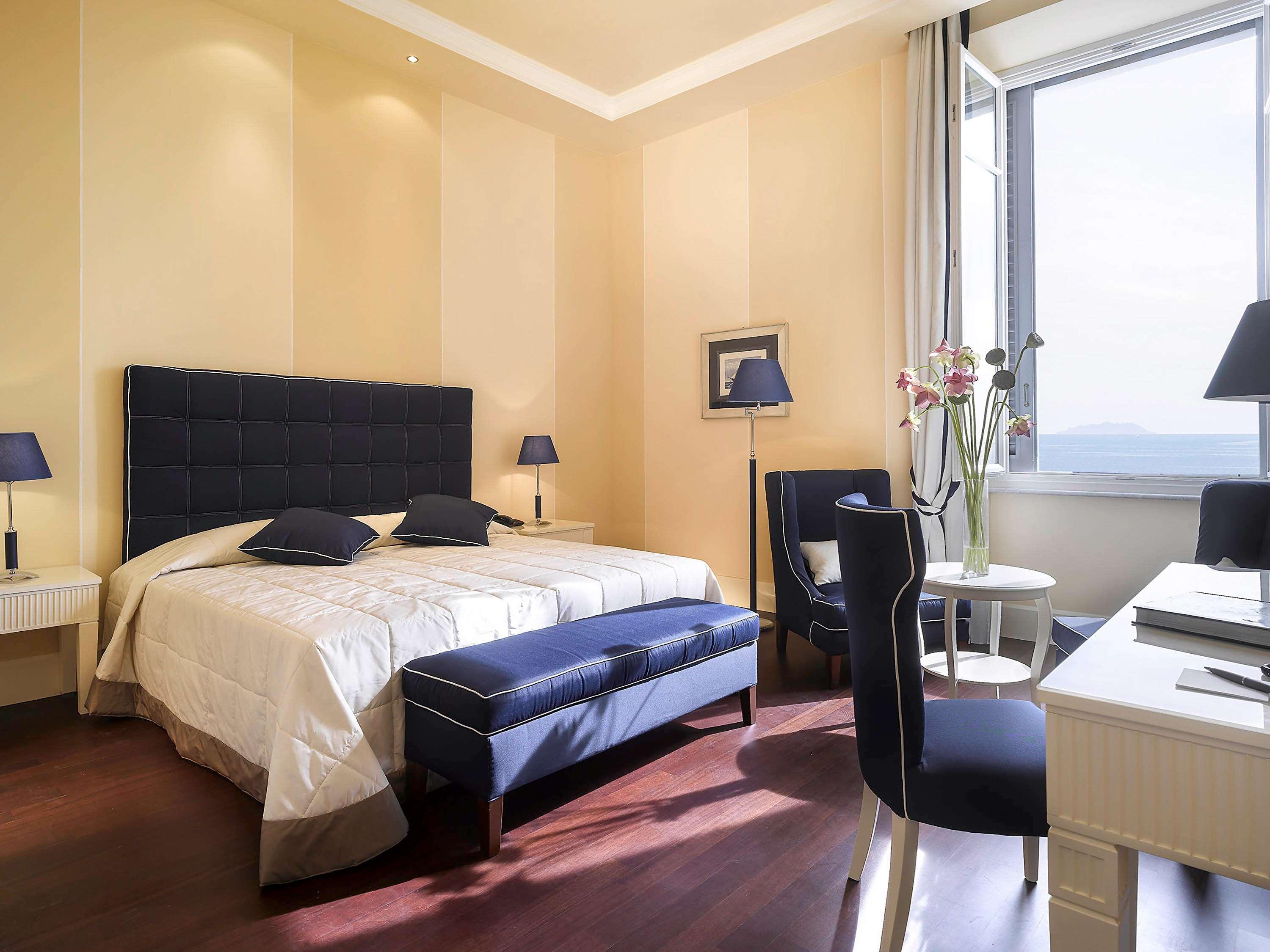 Grand Hotel Palazzo Livorno MGallery Collection Camera foto The photo shows a well-furnished bedroom. It features a large bed with a dark blue headboard and matching pillows, covered with a light-colored bedspread. There are two bedside lamps on either side of the bed. In the room, there is a chair and a smal