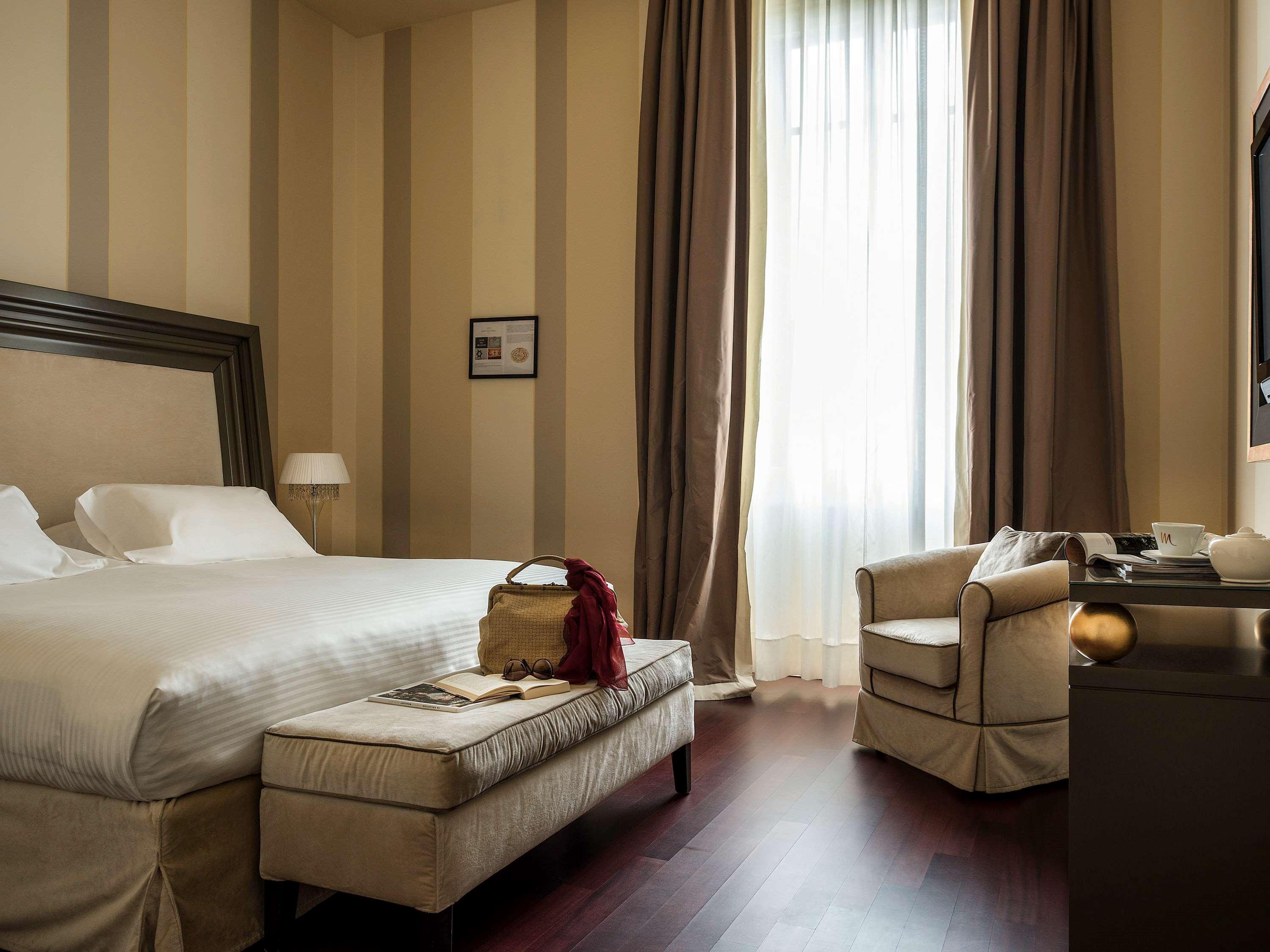 Grand Hotel Palazzo Livorno MGallery Collection Esterno foto The photo shows a elegantly designed hotel room. It features soft, neutral colors with striped walls. There is a large bed with white linens, positioned next to a window draped with long, light-colored curtains that allow natural light to filter in. 