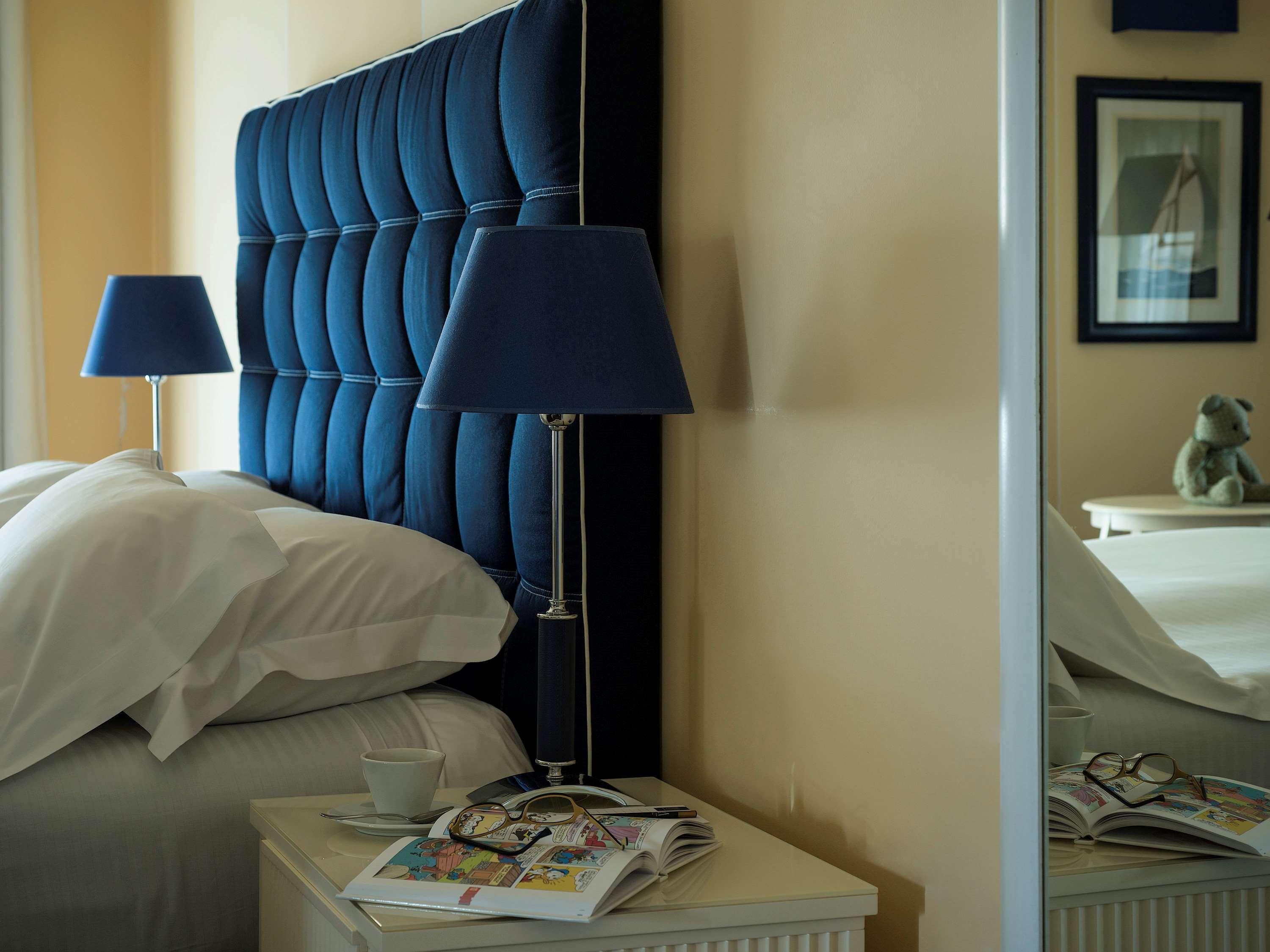Grand Hotel Palazzo Livorno MGallery Collection Esterno foto The photo shows a cozy bedroom setting. There is a large, tufted headboard in a deep blue color behind a neatly made bed with white bedding. On either side of the bed are bedside tables, each featuring a blue lamp. One table has a small cup and a sta