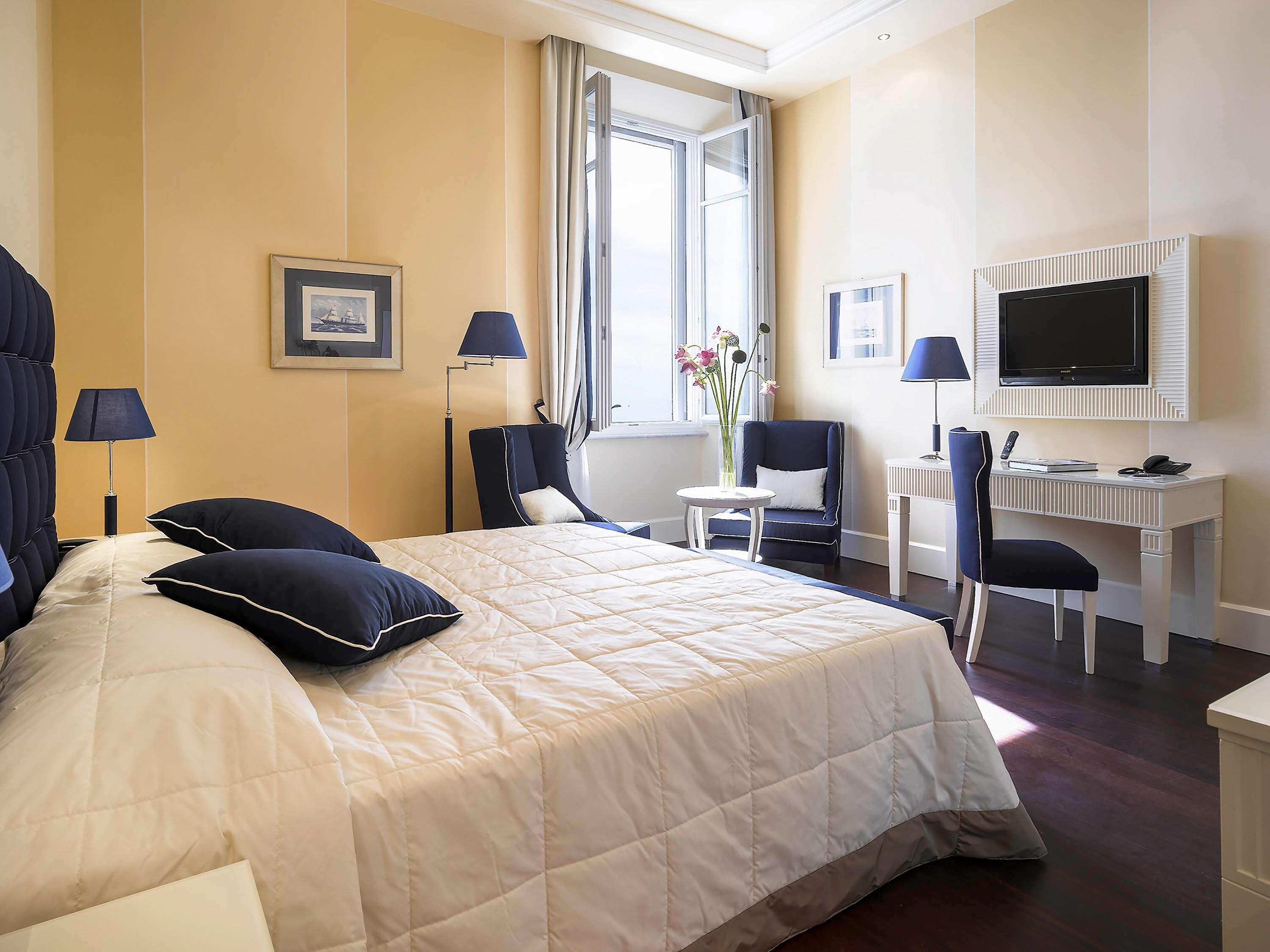 Grand Hotel Palazzo Livorno MGallery Collection Esterno foto The photo shows a well-furnished hotel room. It features a large bed with a white quilt and blue accented pillows. The walls are painted a soft yellow, and there's a window that allows natural light to enter the space. Two blue chairs are positioned 