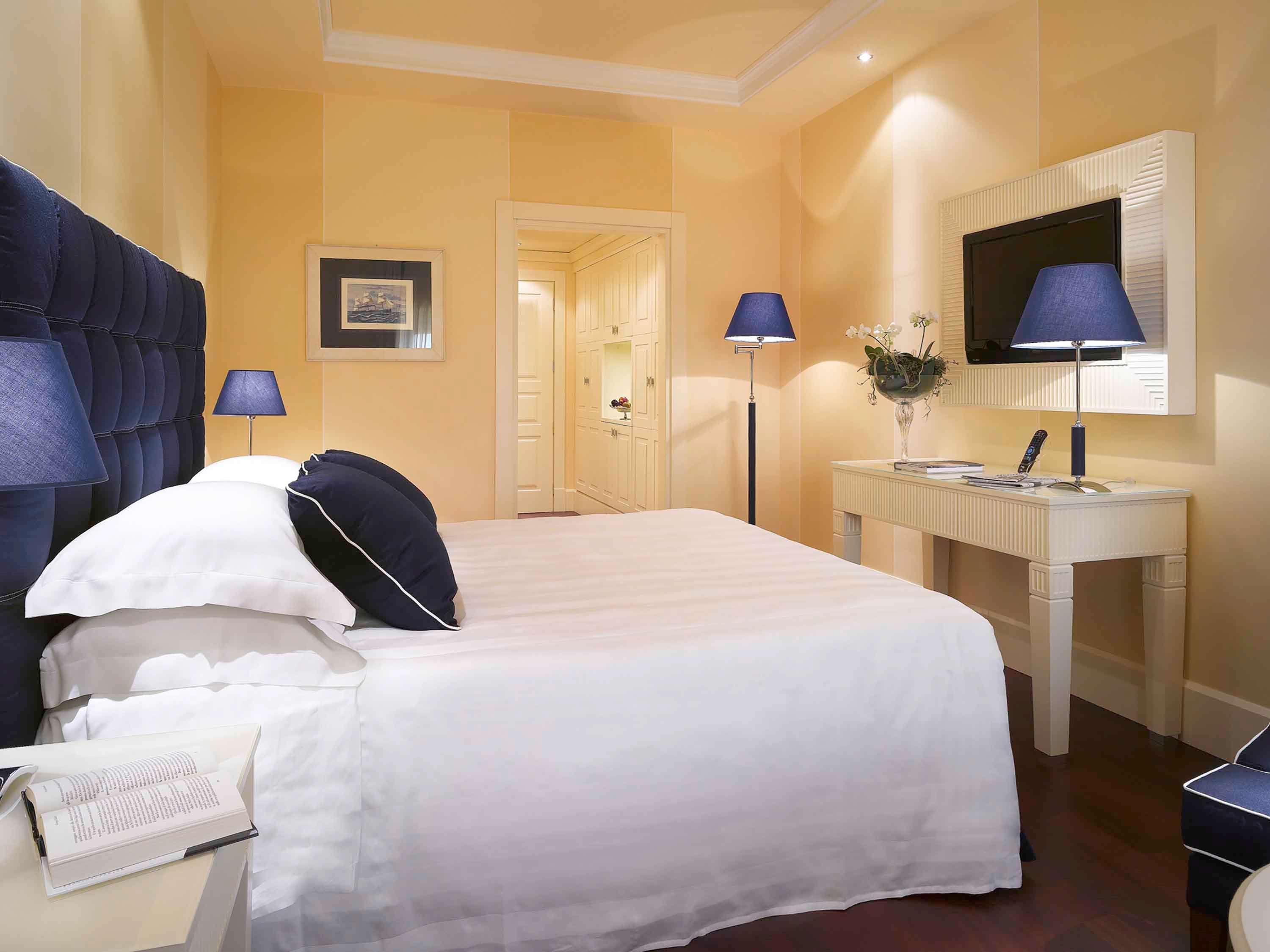 Grand Hotel Palazzo Livorno MGallery Collection Esterno foto The photo shows a well-decorated hotel room featuring a bed with a white coverlet and a dark blue accent on the headboard. There are two bedside lamps with blue shades, and a small table next to the bed. On the wall opposite the bed, there's a flat-s