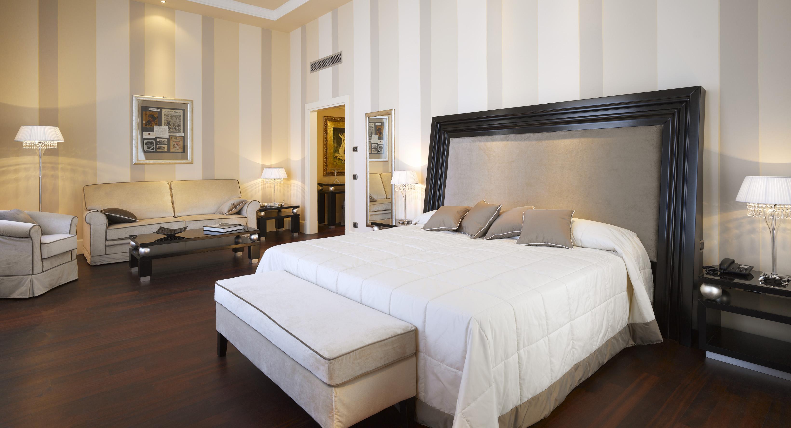 Grand Hotel Palazzo Livorno MGallery Collection Esterno foto The photo shows a stylish hotel room that features a large bed with a light-colored quilt and matching pillows. There is a comfortable seating area to the left, with a beige sofa and a coffee table. On either side of the bed, there are bedside tables