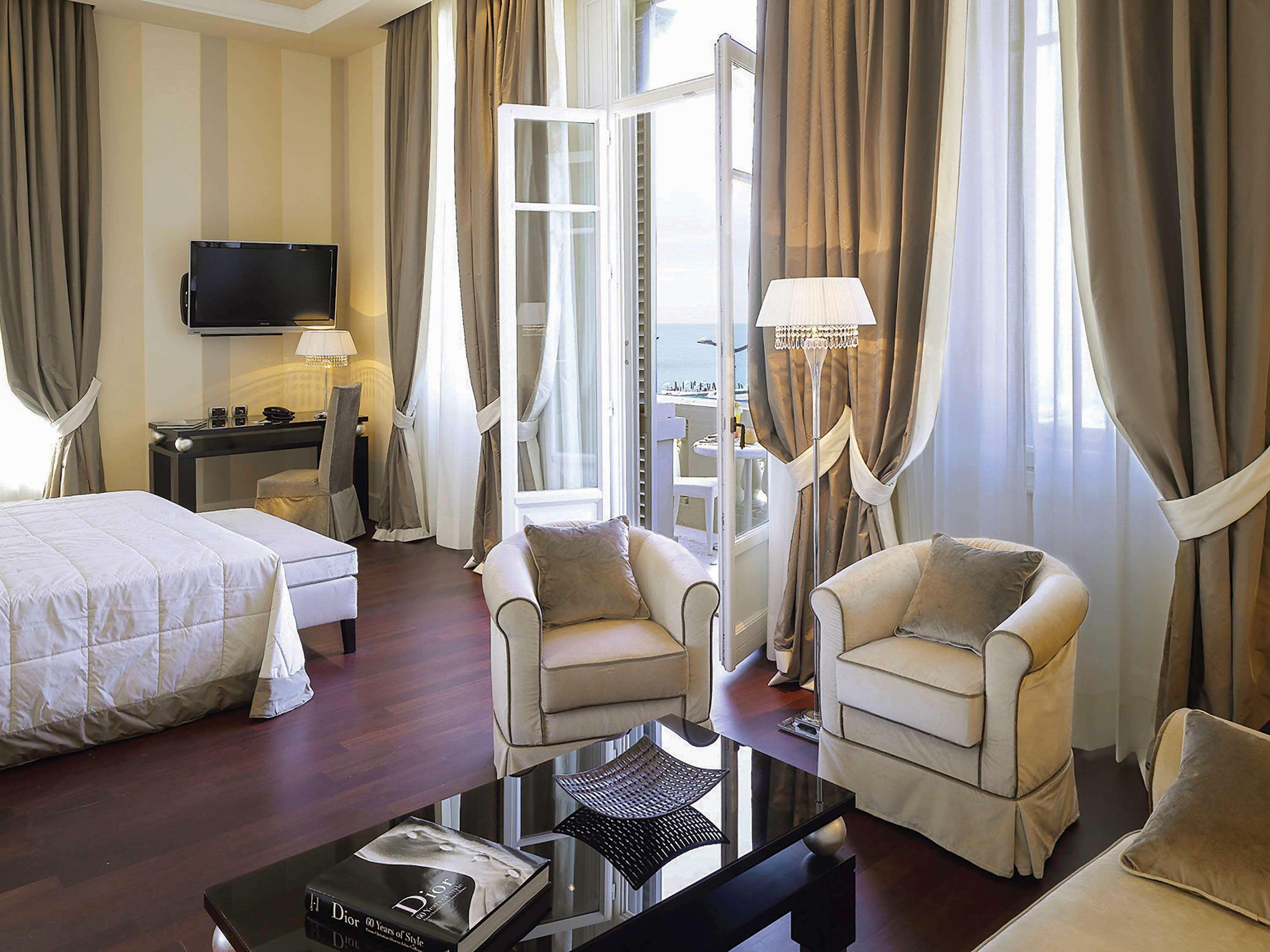 Grand Hotel Palazzo Livorno MGallery Collection Esterno foto The photo shows a well-appointed hotel room or an elegant residential space. It features:

- A large bed with a light-colored quilt and decorative pillows.
- Two comfortable armchairs in a soft, light fabric.
- A sleek black coffee table in the cente