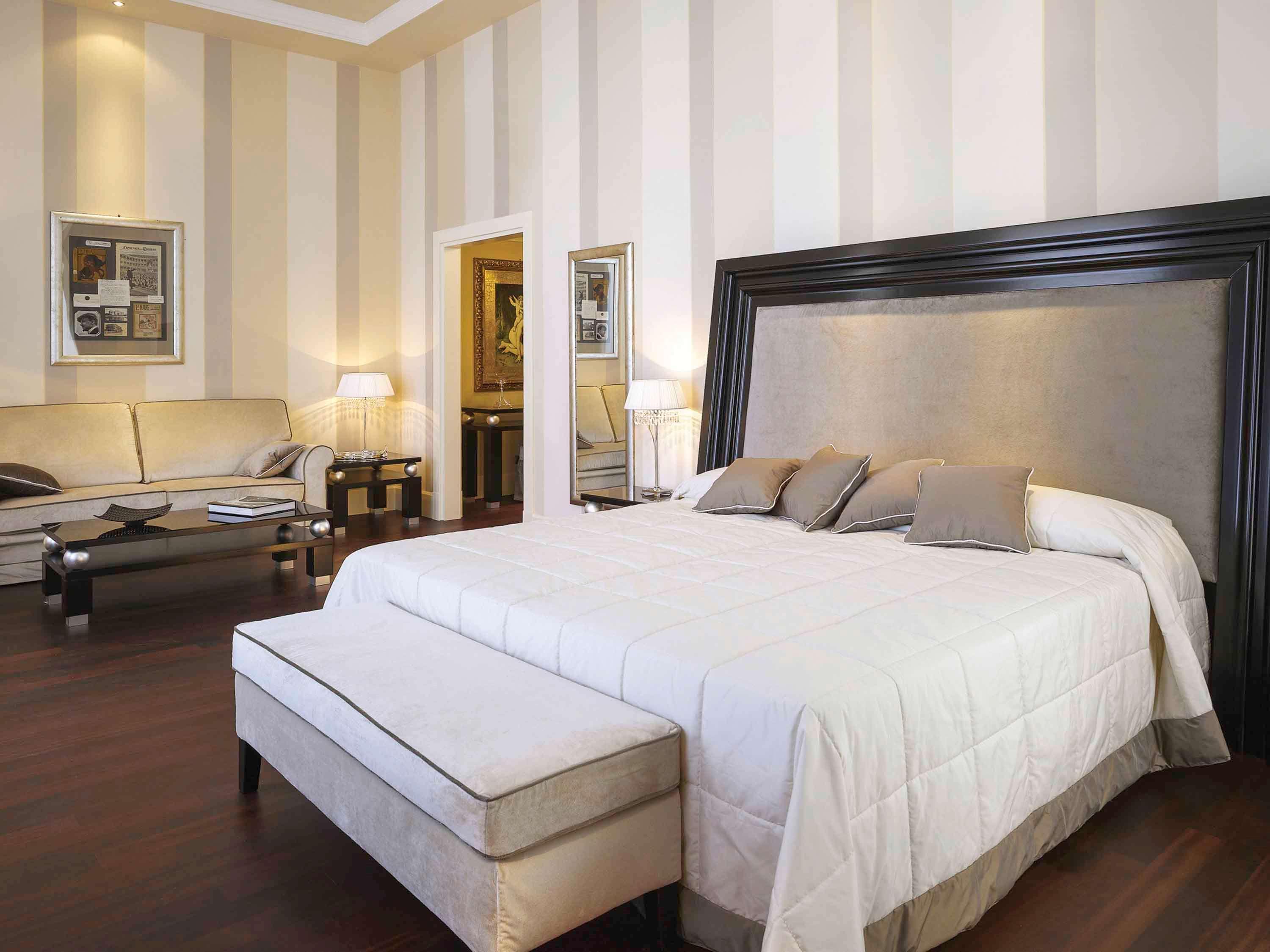 Grand Hotel Palazzo Livorno MGallery Collection Esterno foto The photo shows a stylish bedroom featuring a large, elegant bed with a light-colored quilted comforter and several pillows. At the foot of the bed, there is a padded bench. The room has light-colored striped walls and dark hardwood flooring. To the 