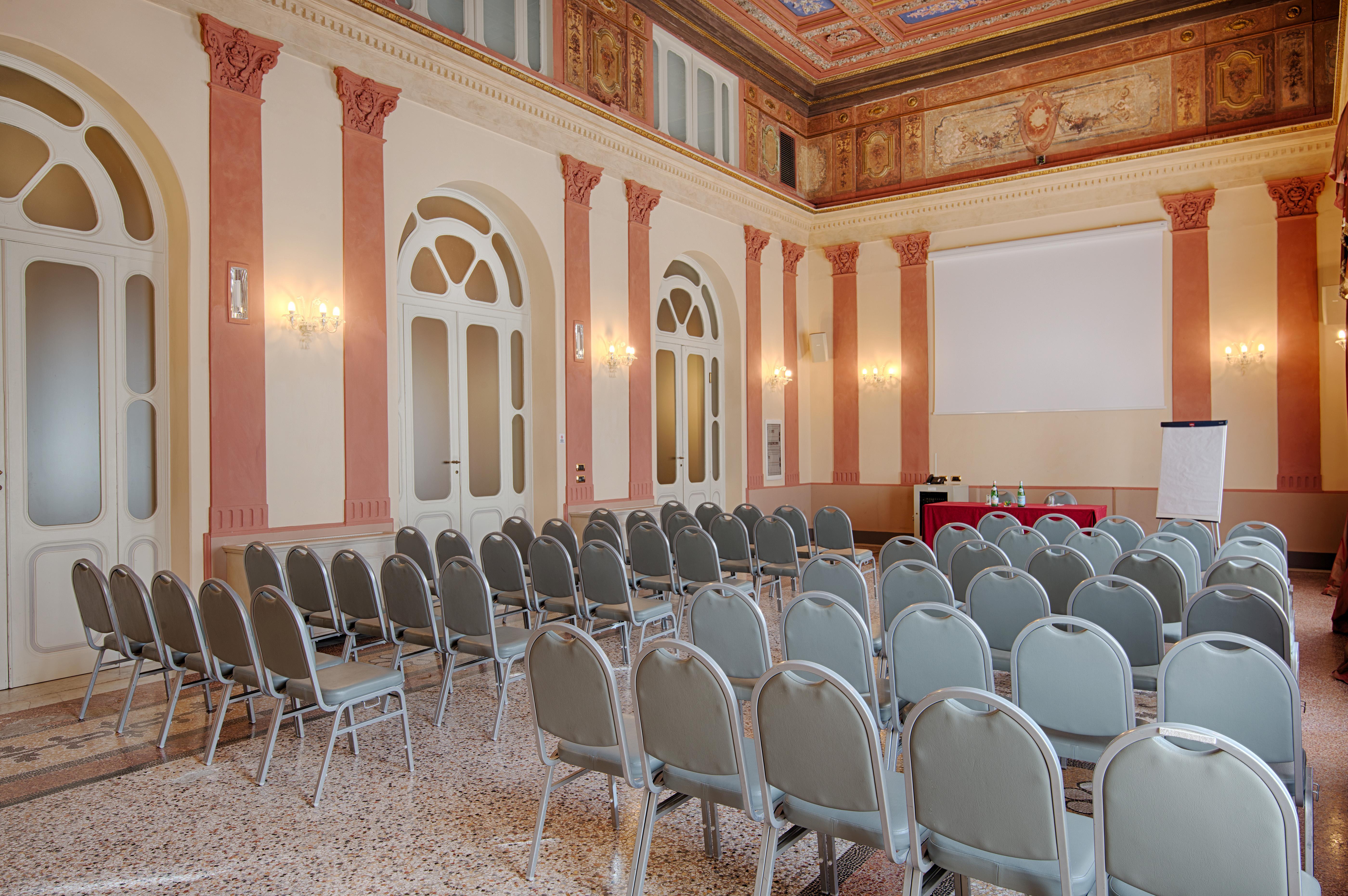 Grand Hotel Palazzo Livorno MGallery Collection Esterno foto The photo shows a spacious room set up for an event, likely a conference or meeting. There are several rows of gray chairs arranged facing a blank wall, where a projector screen may be placed. The room features elegant architectural details with ligh