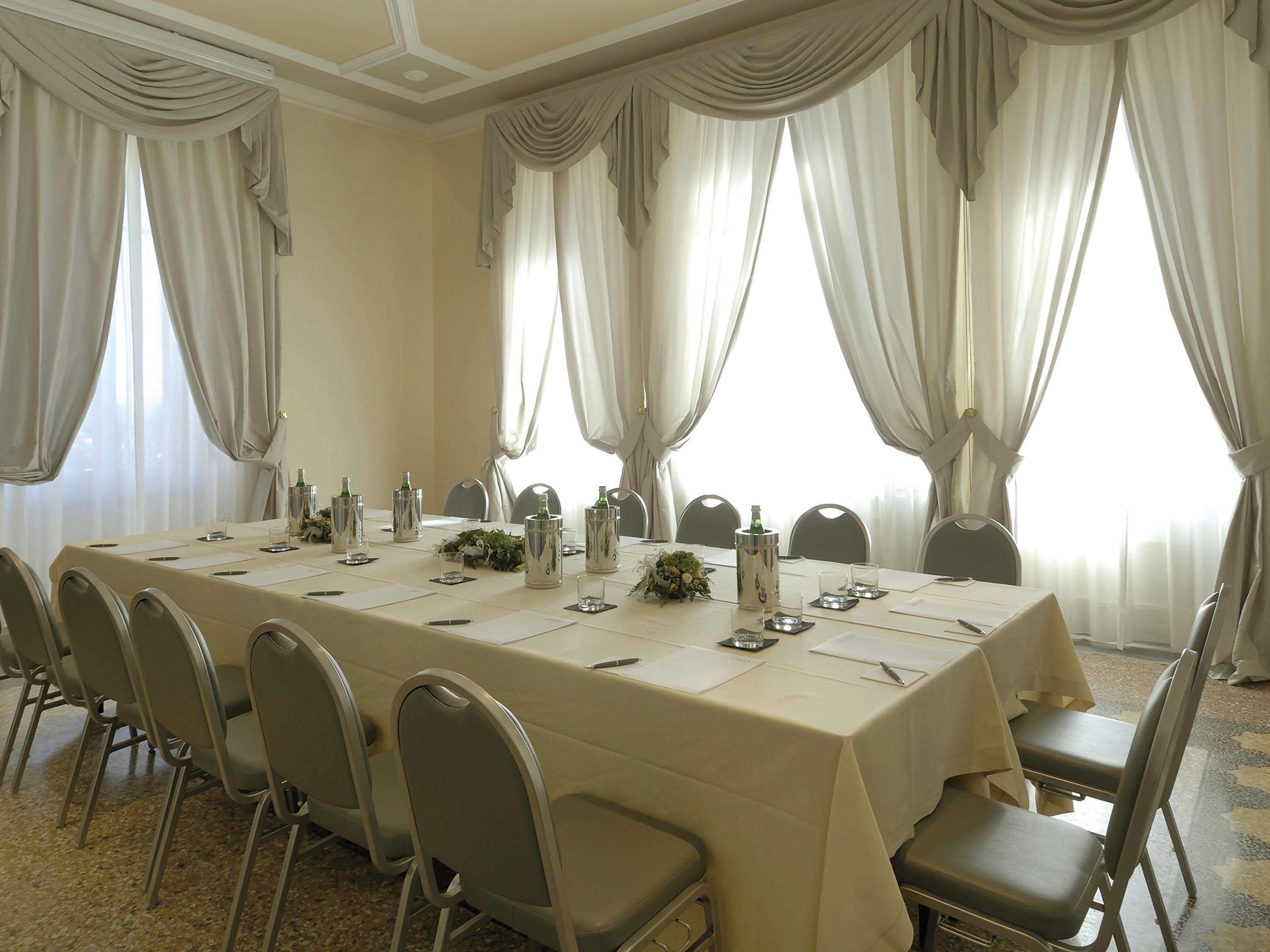 Grand Hotel Palazzo Livorno MGallery Collection Esterno foto The photo shows a well-lit meeting room with large windows draped with elegant curtains. In the center, there is a long table covered with a light-colored tablecloth. The table is set with several water bottles and neatly arranged utensils. Around th