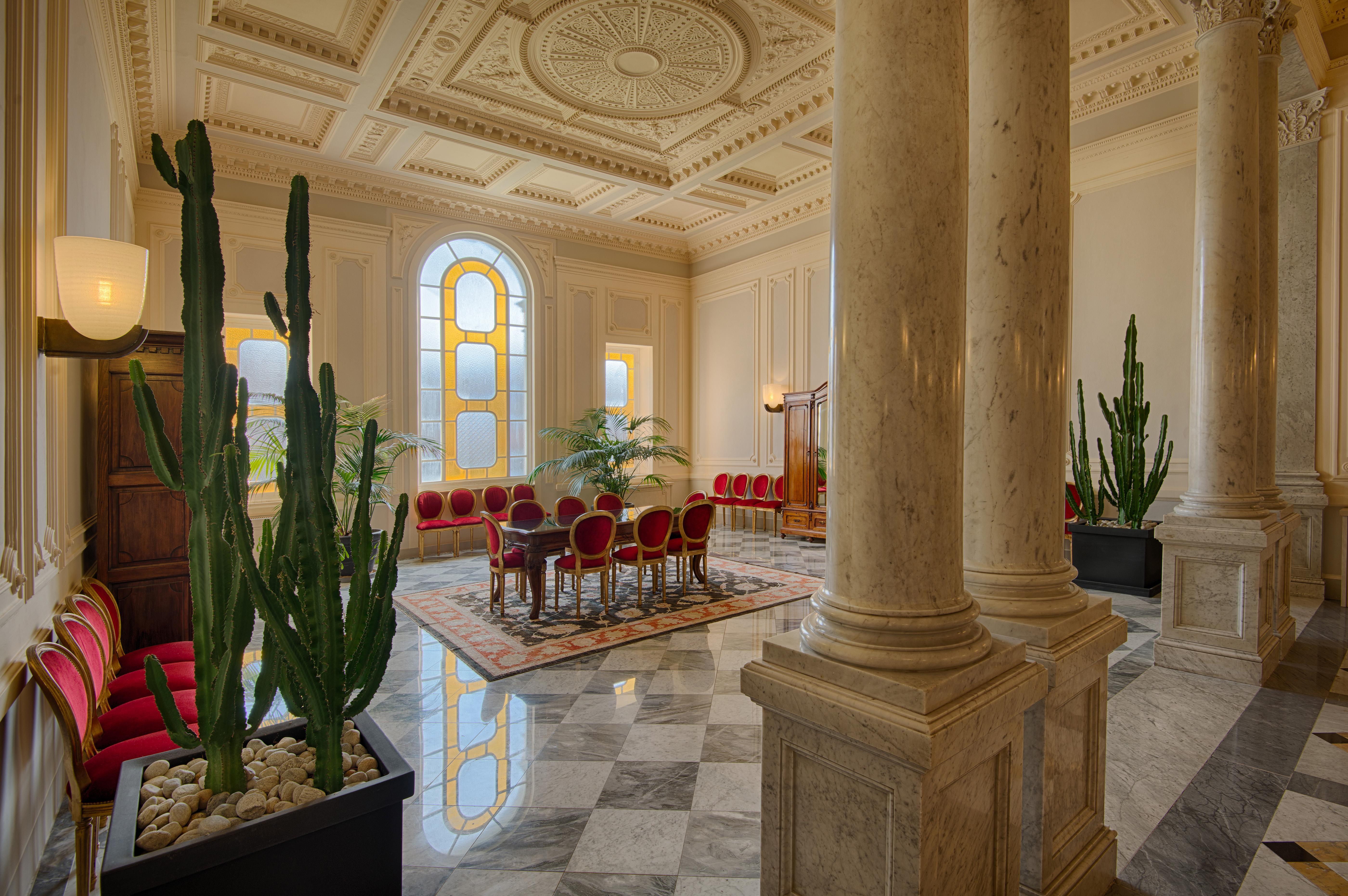 Grand Hotel Palazzo Livorno MGallery Collection Esterno foto The photo shows an elegant interior space, likely a waiting room or meeting area. The room features tall columns and a high, decorative ceiling with intricate designs. There are red upholstered chairs arranged in a semicircle around a table in the ce
