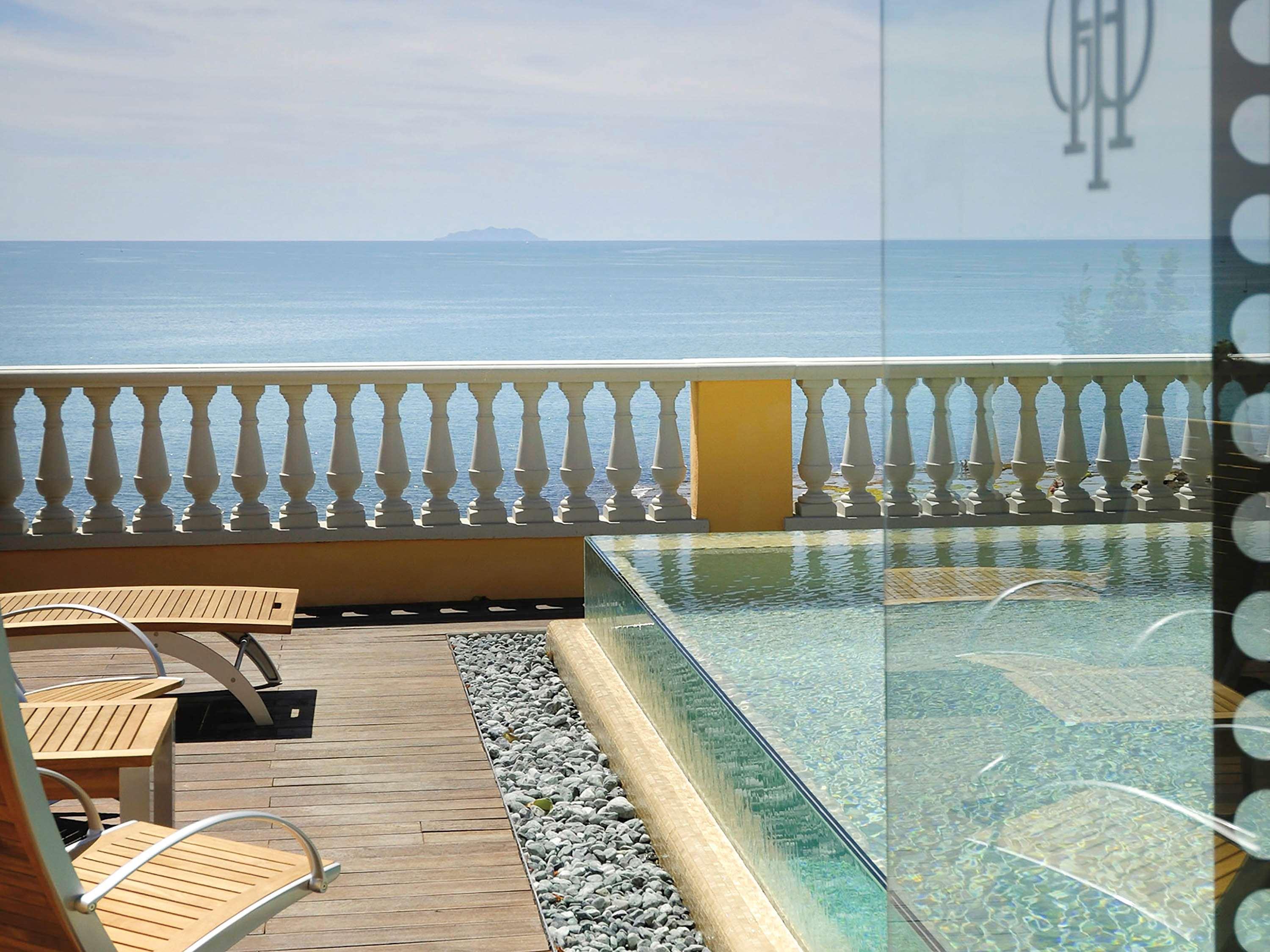 Grand Hotel Palazzo Livorno MGallery Collection Esterno foto The photo showcases a serene view of a poolside area with an infinity pool that appears to blend into the ocean beyond. There are wooden loungers positioned on a wooden deck next to the pool. A decorative railing can be seen in the foreground, provid