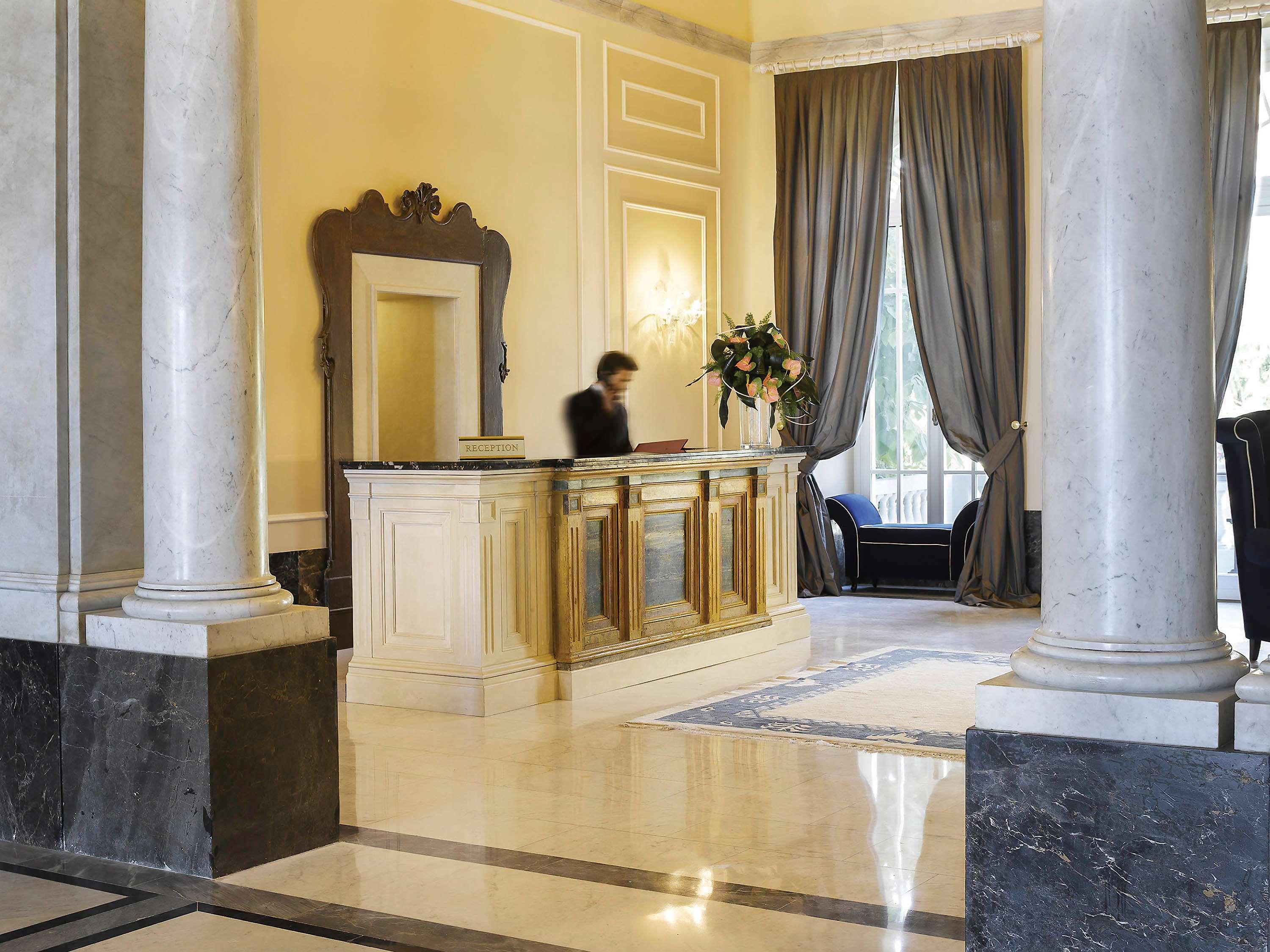 Grand Hotel Palazzo Livorno MGallery Collection Esterno foto The photo shows a hotel lobby area. In the foreground, there is a reception desk made of wood with elegant design elements. Behind the desk, a staff member is engaged in their duties. The lobby features tall, light-colored walls and large windows dre