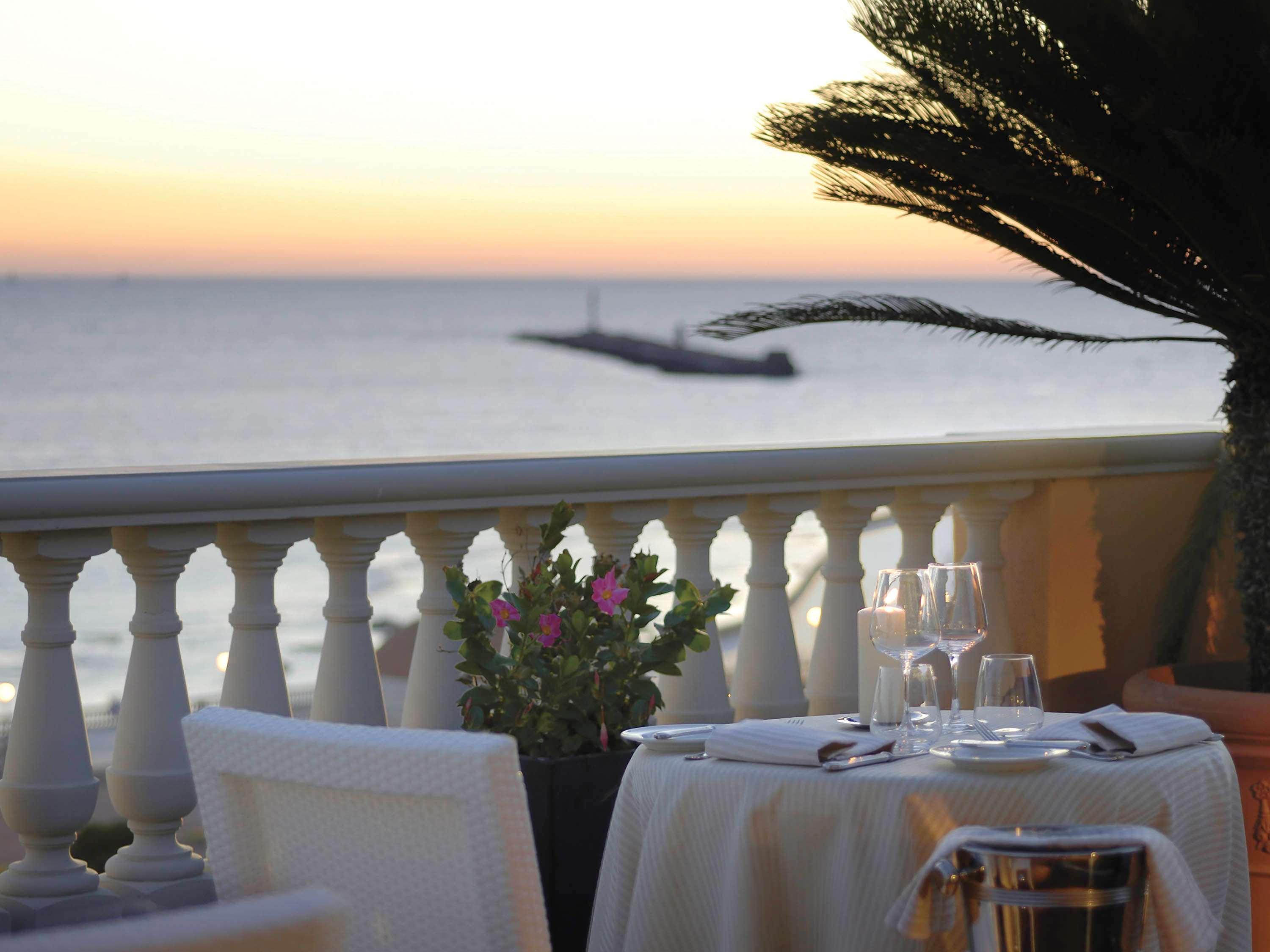 Grand Hotel Palazzo Livorno MGallery Collection Esterno foto The photo shows a picturesque dining setting on a balcony overlooking the sea at sunset. There is a elegantly set table with a white tablecloth, glassware, and a potted plant with pink flowers as a centerpiece. In the background, the calm sea reflect