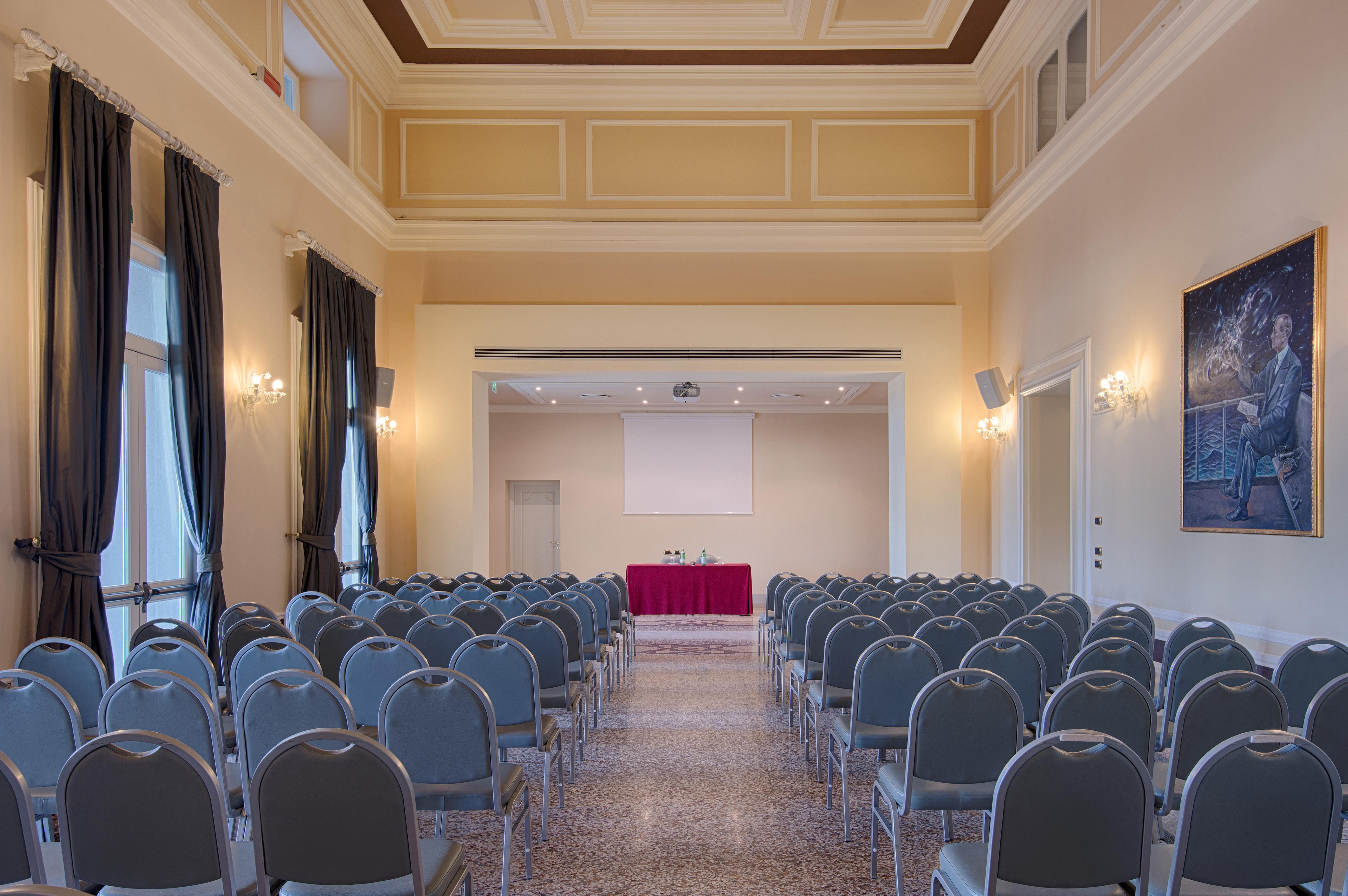 Grand Hotel Palazzo Livorno MGallery Collection Esterno foto The photo depicts a conference or meeting room set up with rows of gray chairs facing a stage at the front. The room features high ceilings with decorative moldings and large windows draped with dark curtains. A red table is positioned at the front o