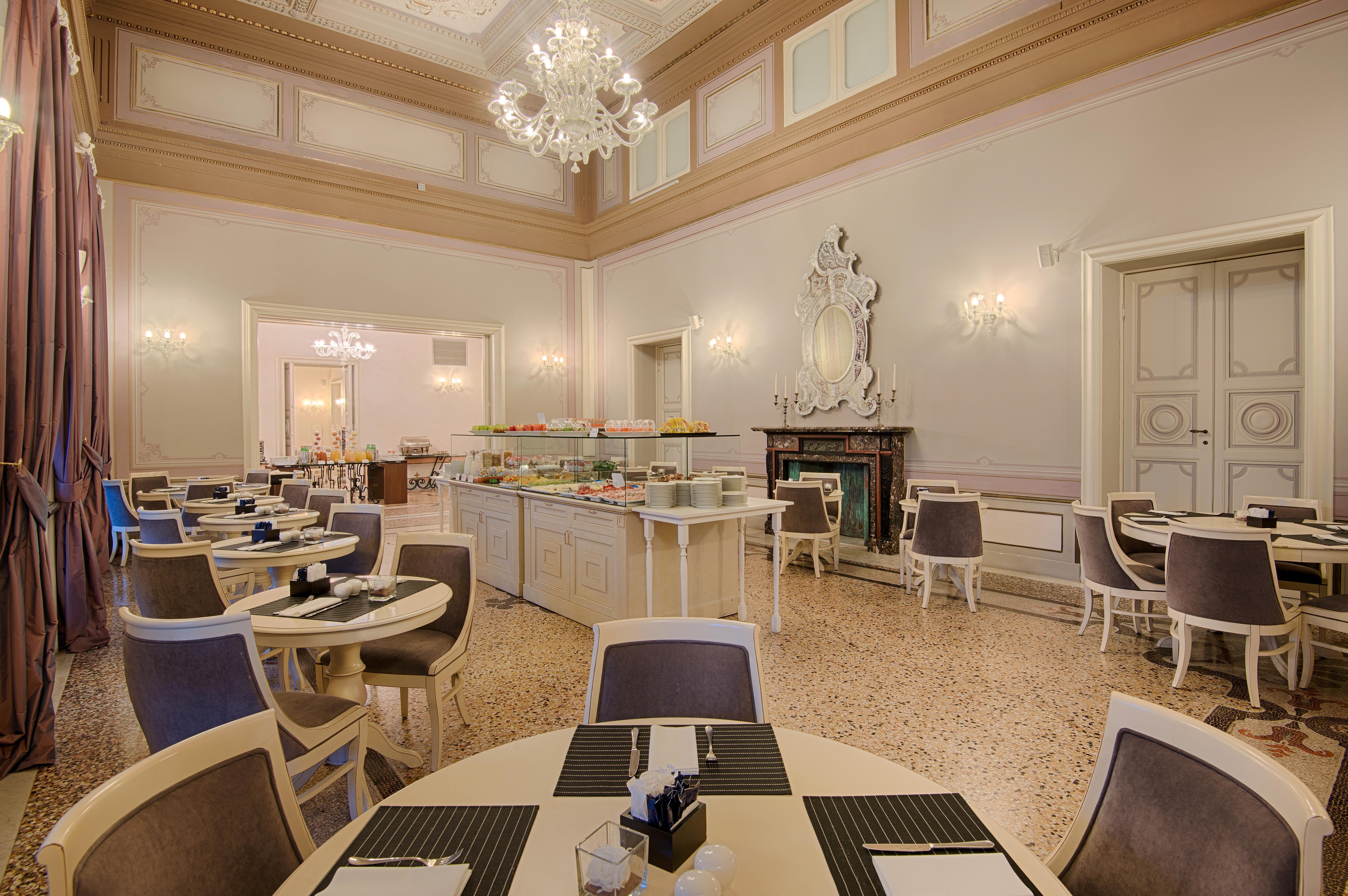 Grand Hotel Palazzo Livorno MGallery Collection Esterno foto The photo depicts an elegant restaurant interior. It features light-colored walls with ornate moldings and a high ceiling adorned with a chandelier. The room is arranged with several round tables, each set with black place mats and tableware. A buffe