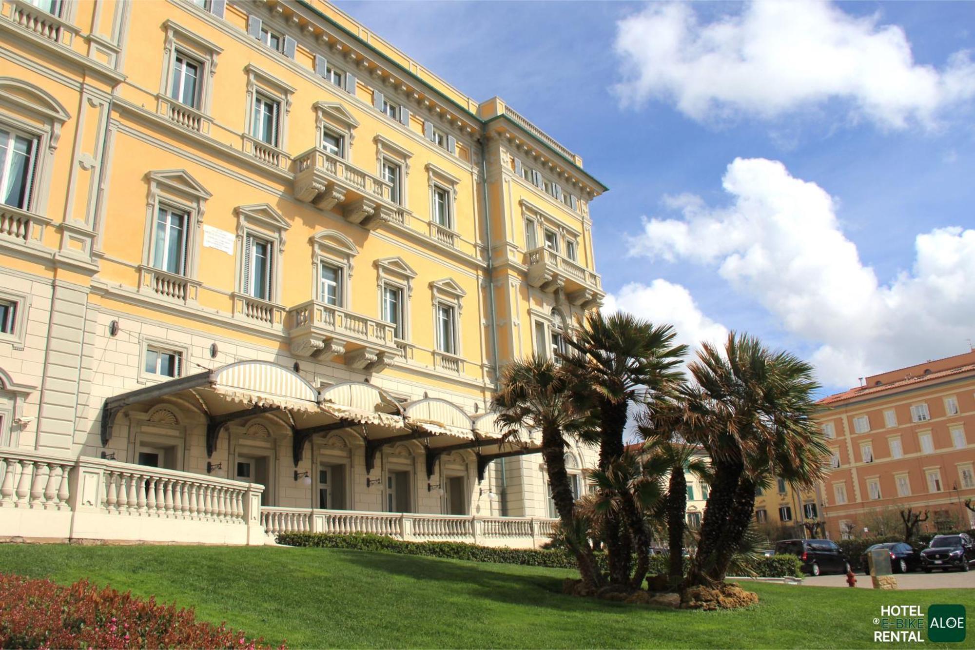 Grand Hotel Palazzo Livorno MGallery Collection Esterno foto The image shows a large, elegant building with a light-yellow exterior. The architecture features multiple windows with decorative elements. In the foreground, there is a well-maintained lawn with greenery and palm trees. The sky is partly cloudy, in